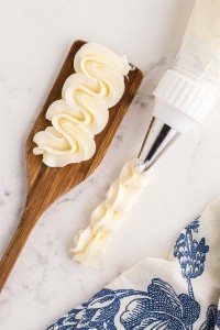 Piping bag filled with Italian meringue buttercream, wooden board with piped buttercream, blue and white decorative linen, on a marble countertop