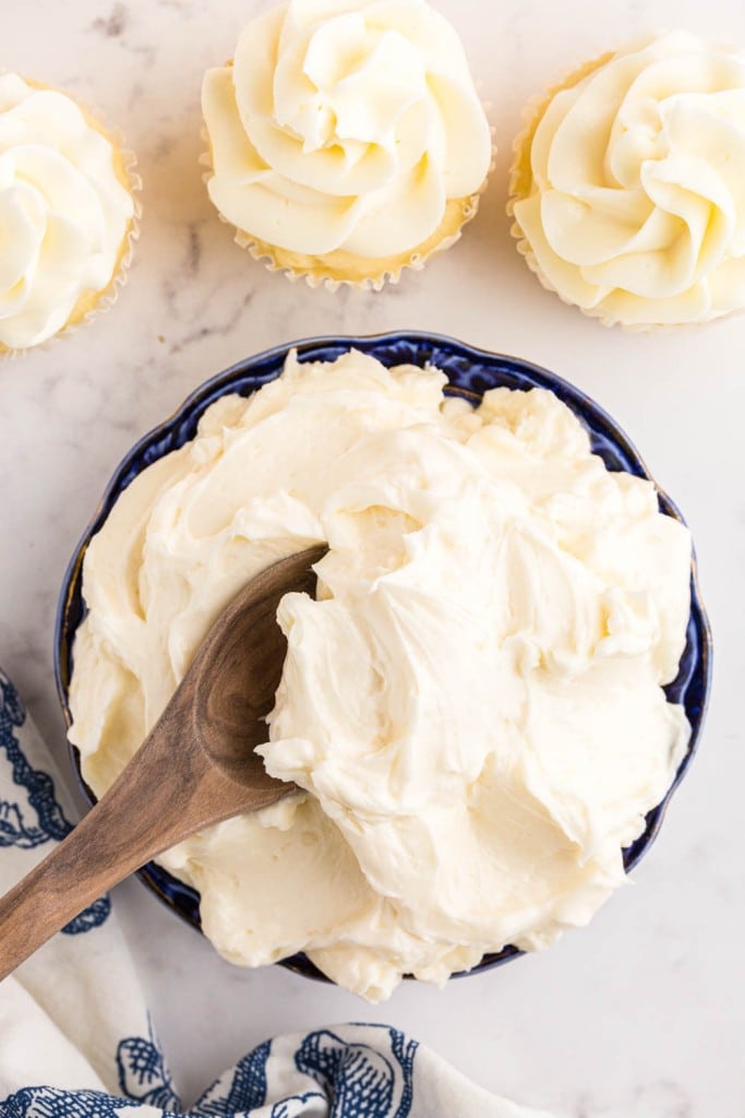 Bowl of Italian meringue buttercream, wooden mixing spoon submerged in bowl of buttercream, three frosted vanilla cupcakes, blue and white decorative linen, on a marble countertop