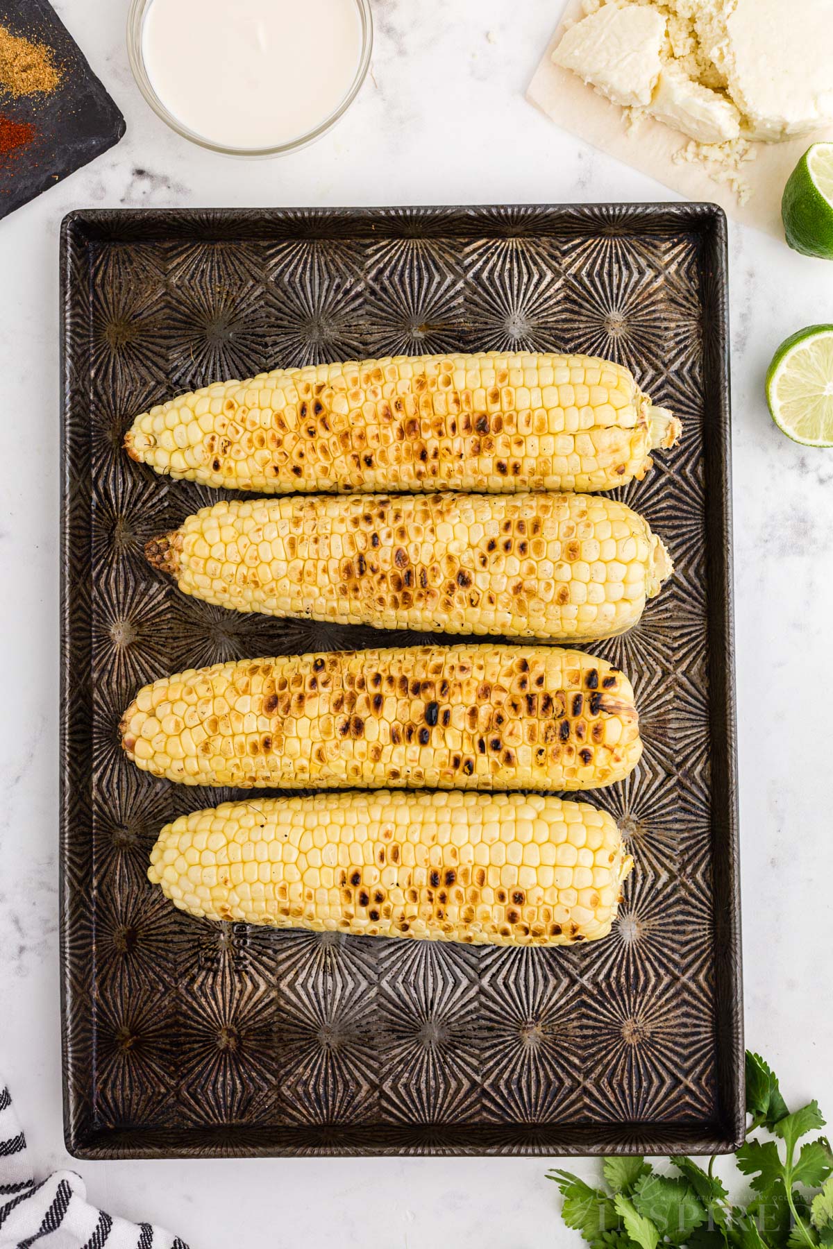 Ears of corn with charred marks on baking sheet, Mexican street corn sauce ingredients set nearby.