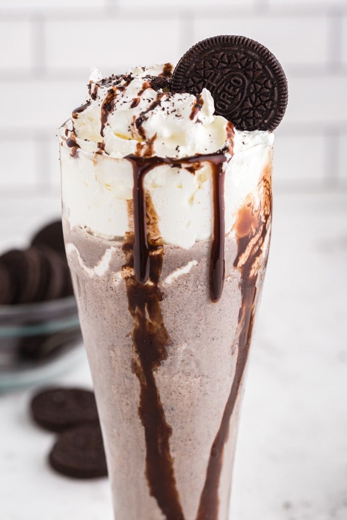 Oreo milkshake in a tall glass decorated with whipped cream, chocolate syrup, and an Oreo cookie, bowl of Oreo cookies, on marble countertop, white stacked tiles in the background