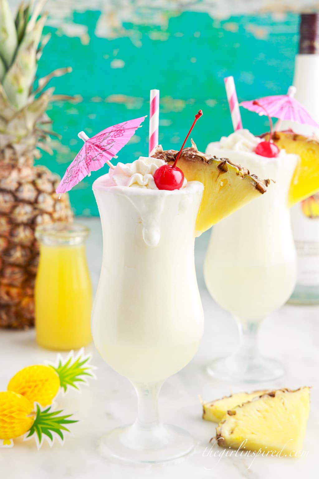 two pina coladas next to a whole pineapple and a glass bottle of pineapple juice