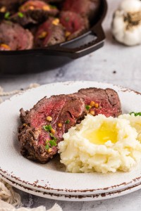 White plate of roasted beef tenderloin with a portion of mashed potatoes, baking dish with roasted beef tenderloin, whole clove of garlic, kitchen towel, on a marble surface