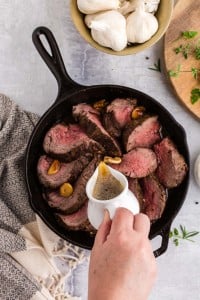 Pouring the garlic browned butter over the sliced roasted beef tenderloin, kitchen towel, bowl of garlic cloves, fresh herbs on wooden board, on top of a marble surface