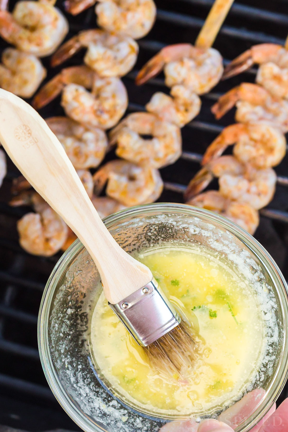Bowl of garlic butter sauce and basting brush held over grill with shrimp skewers on it.