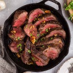 Roasted Beef Tenderloin in a skillet, kitchen towel, plate with forks, fresh herbs on a wooden board, on a marble surface