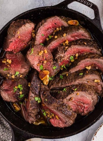Roasted Beef Tenderloin in a skillet, kitchen towel, plate with forks, fresh herbs on a wooden board, on a marble surface