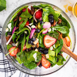 Glass bowl filled with tossed Strawberry Feta Spinach Salad, bowl with strawberries, half an avocado, bowl with blueberries, bowl with onions, black and white checked linen on a marble countertop.