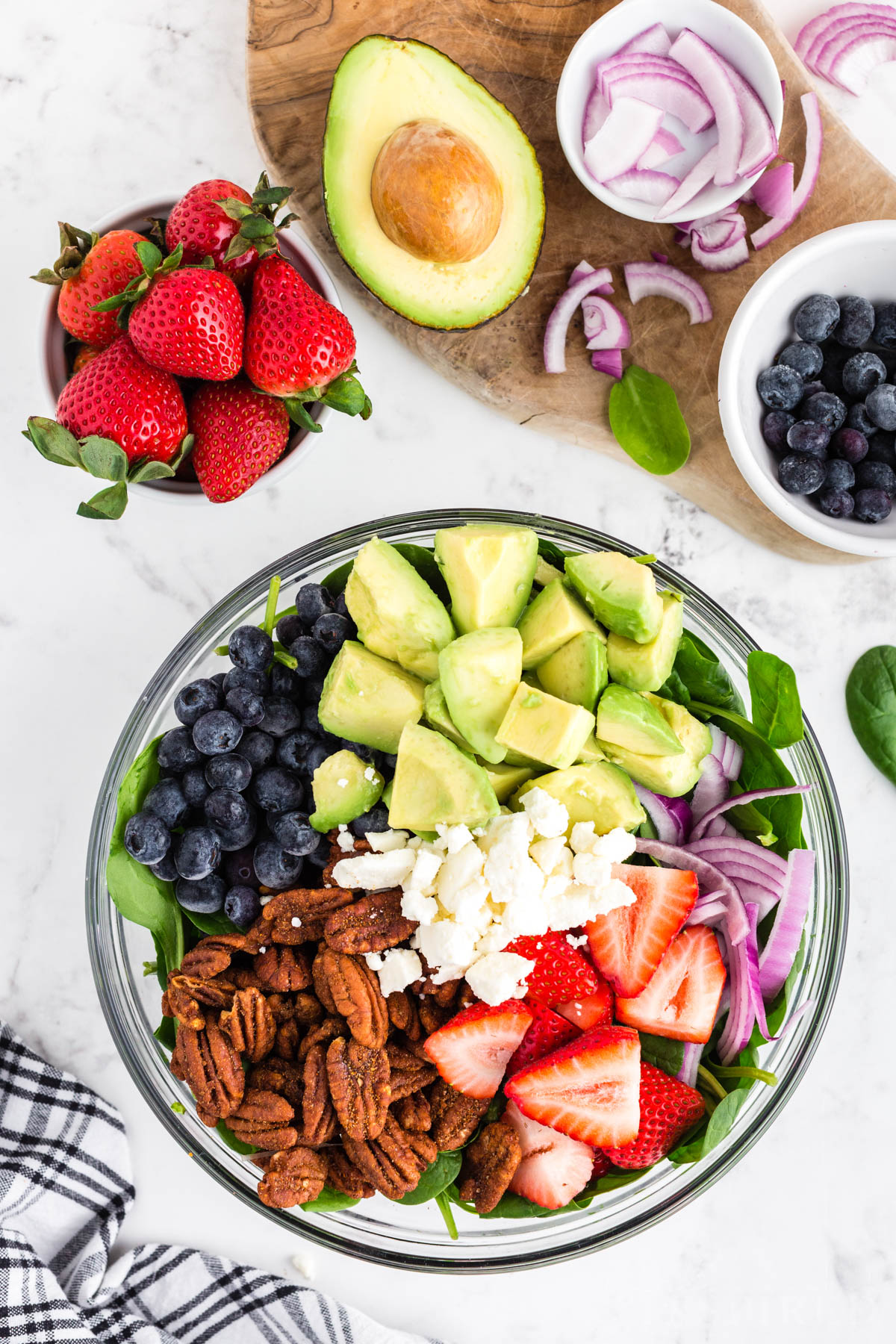 Glass bowl filled with spinach, pecans, cut strawberries, cut avocado, blueberries, onions, feta cheese, bowl with strawberries, half an avocado, bowl with blueberries, bowl with onions, black and white checked linen on a white marble countertop.