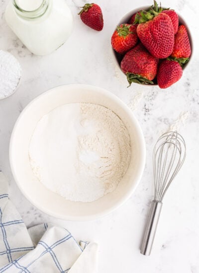 Dry ingredients in mixing bowl with jug of milk and bowl of strawberries.
