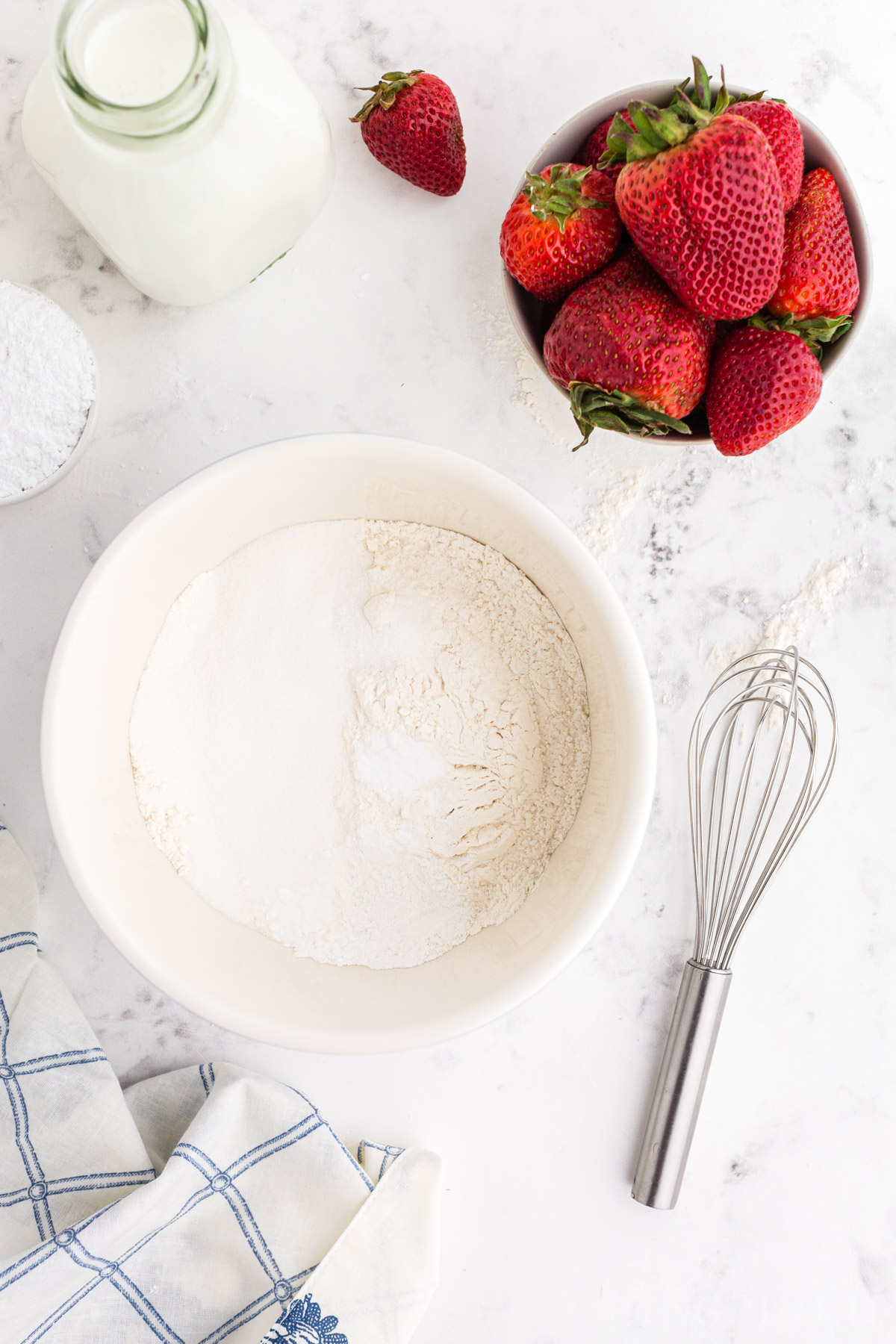 Dry ingredients in mixing bowl with jug of milk and bowl of strawberries.