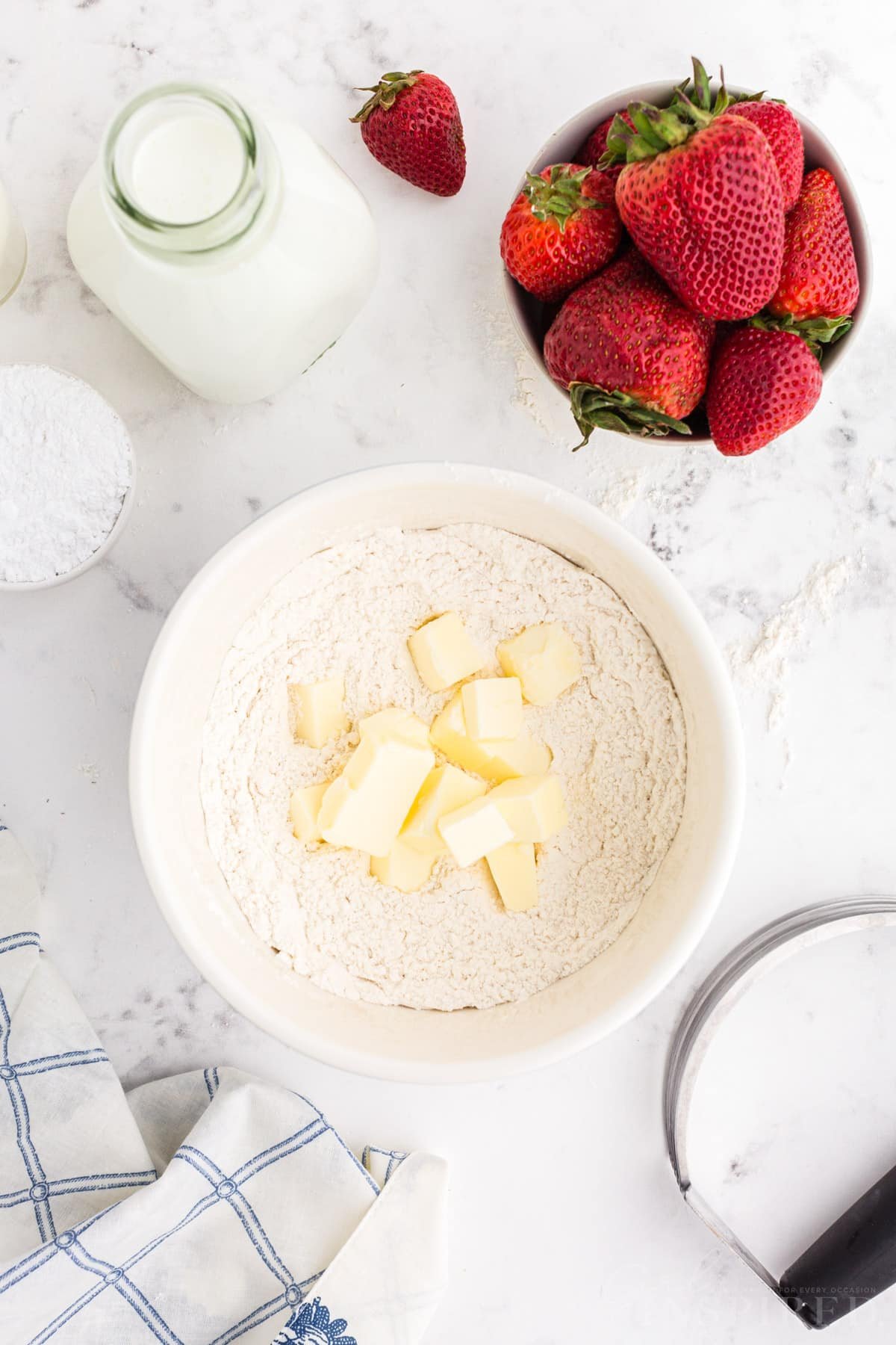 Pieces of butter in dry ingredient mixture with jug of milk and bowl of strawberries.