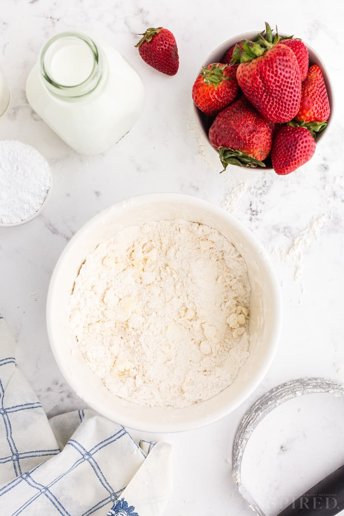 Strawberry scone dry mixture of crumbly ingredients in bowl with strawberries and jar of milk.