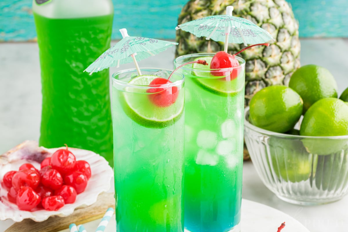 Two glasses of tipsy mermaid rum punch garnished with fruit and a paper umbrella next to bowl of limes, seashell filled with maraschino cherries, a bottle of Midori and a pineapple.