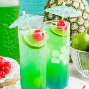 Two tall glasses with layered tipsy mermaid drink, topped with lime slice, maraschino cherry, and paper umbrella, bowl of limes and pineapple in background.