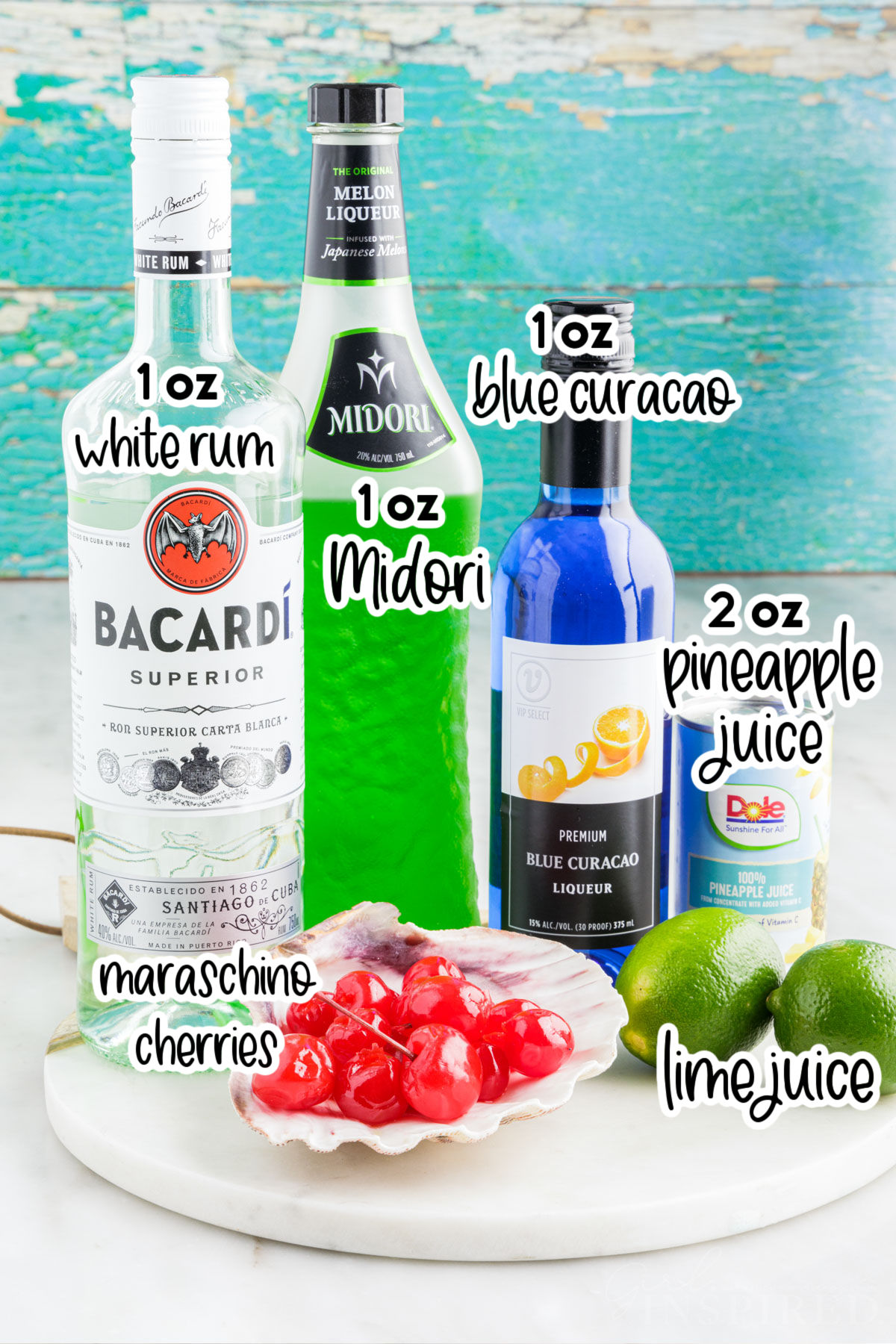 Ingredients to make a tipsy mermaid drink including a bottle of Bacardi rum, Midori, blue curacao, a can of pineapple juice, dish of maraschino cherries, and two limes.