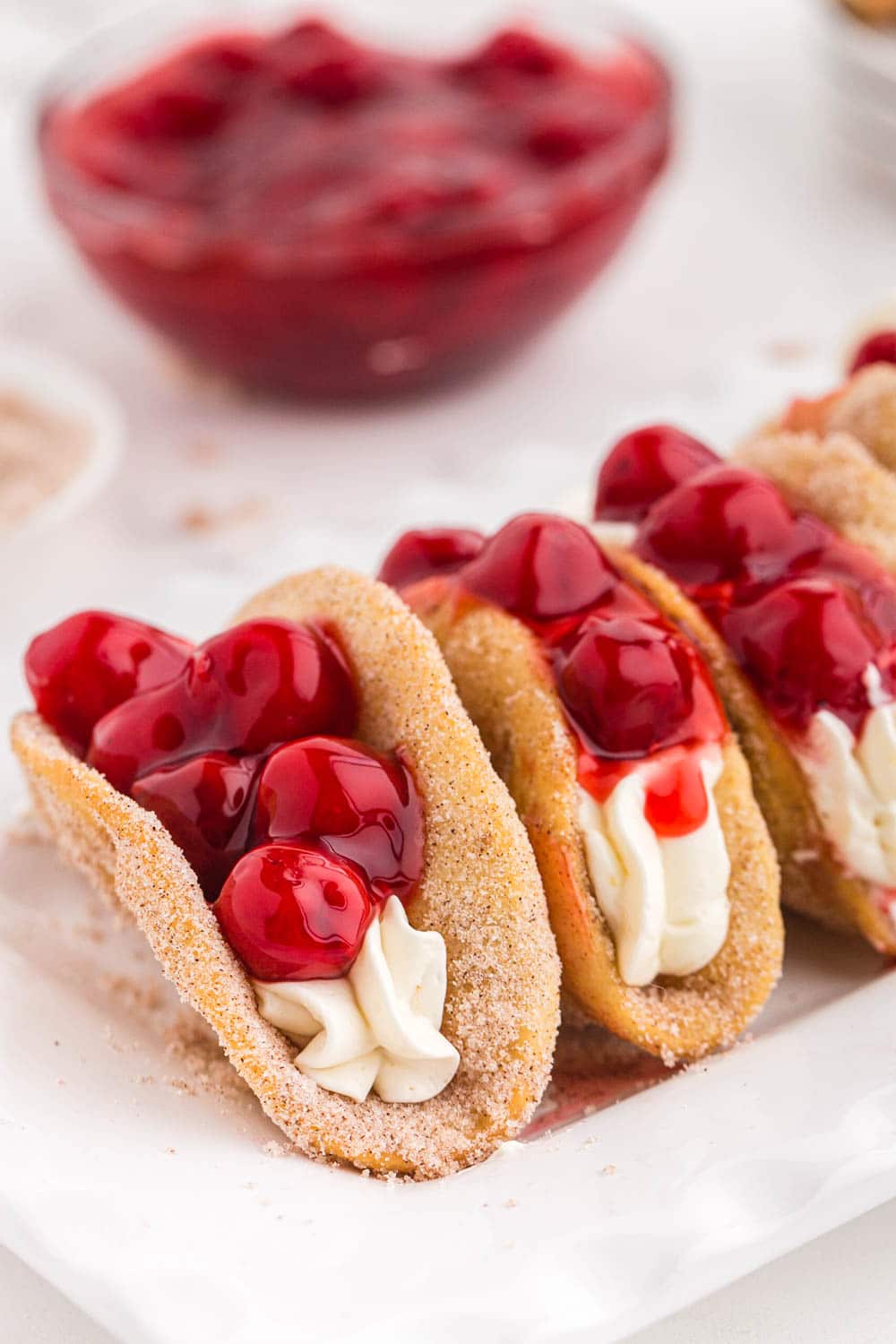 Three cherry cheesecake tacos on a white serving plate, bowl of cherry pie filling on a marble countertop