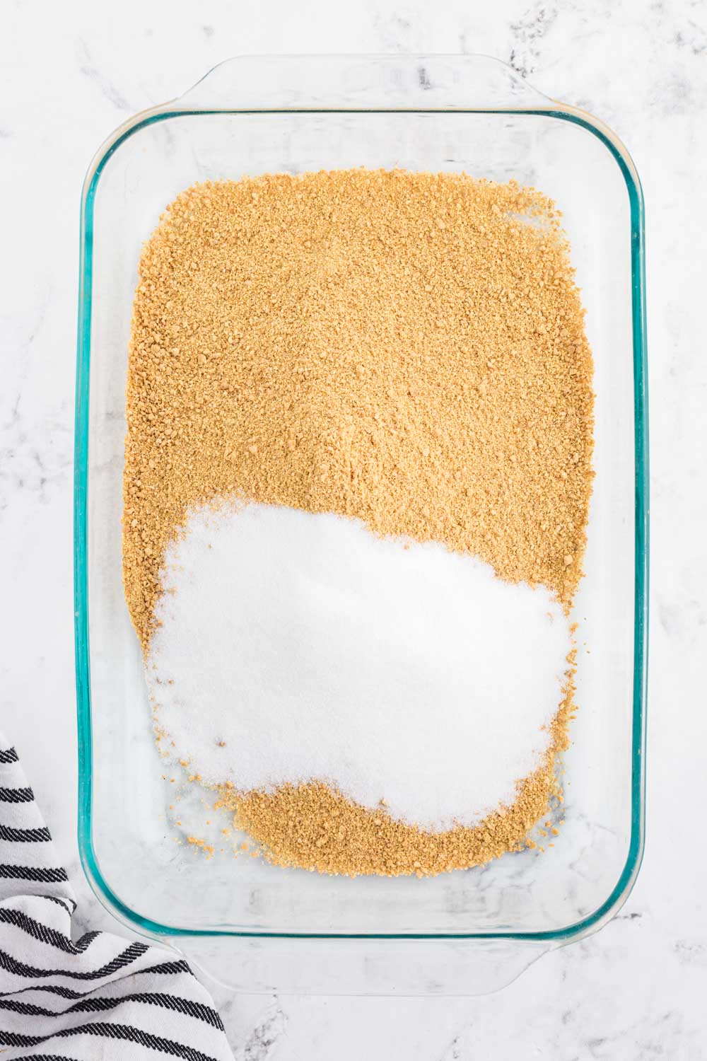 Graham cracker crumbs and granulated sugar added to a 9x13-inch baking dish, striped linen, on a white marble surface.