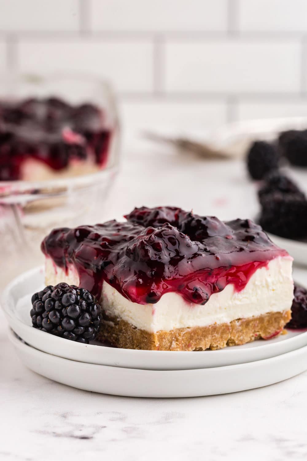 Slice of no-bake blackberry cheesecake on white cake plates, baking dish with blackberry cheesecake, bowl of fresh blackberries, on a white marble surface with white stacked tiles in the background.