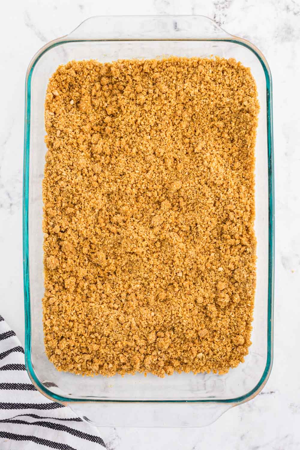 Graham cracker crust mixture added to a 9x13-inch baking dish, striped linen, on a white marble surface.