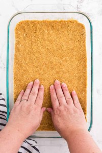 Graham cracker crust mixture pressed with fingers into a 9x13-inch baking dish, striped linen, on a white marble surface.