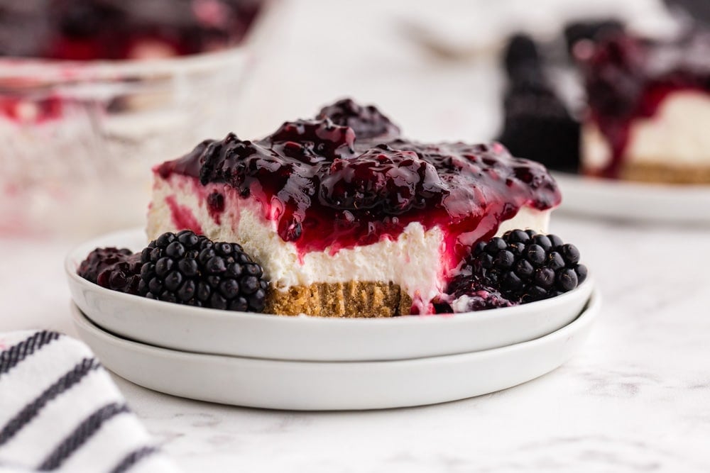 Slice of no bake blackberry cheesecake on cake plate, striped linen, baking dish with no bake blackberry cheesecake, slice of cheesecake in the background, on a white marble countertop.