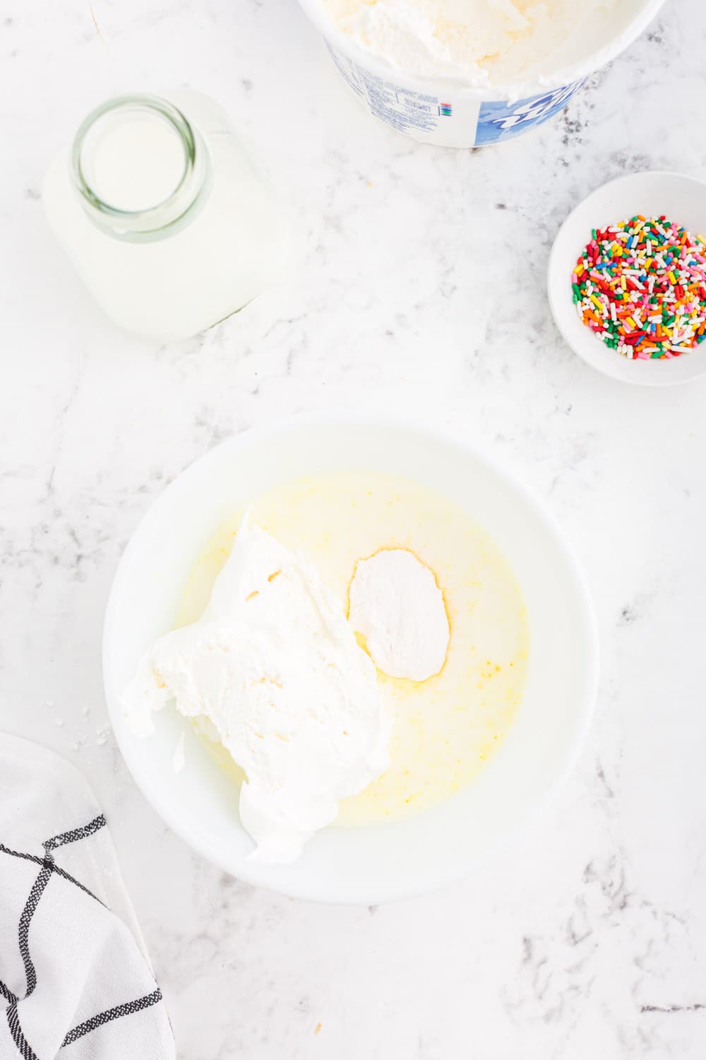 Mixing bowl with vanilla instant pudding mix, milk, and whipped topping, small bowl of rainbow sprinkles, glass jar of milk, striped linen, on a white marble surface.