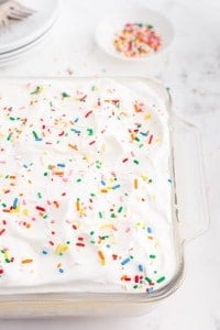 Glass dish with fully assembled birthday cake cheesecake bars, small bowl of rainbow sprinkles, stacked white serving plates with forks, on a white marble surface.