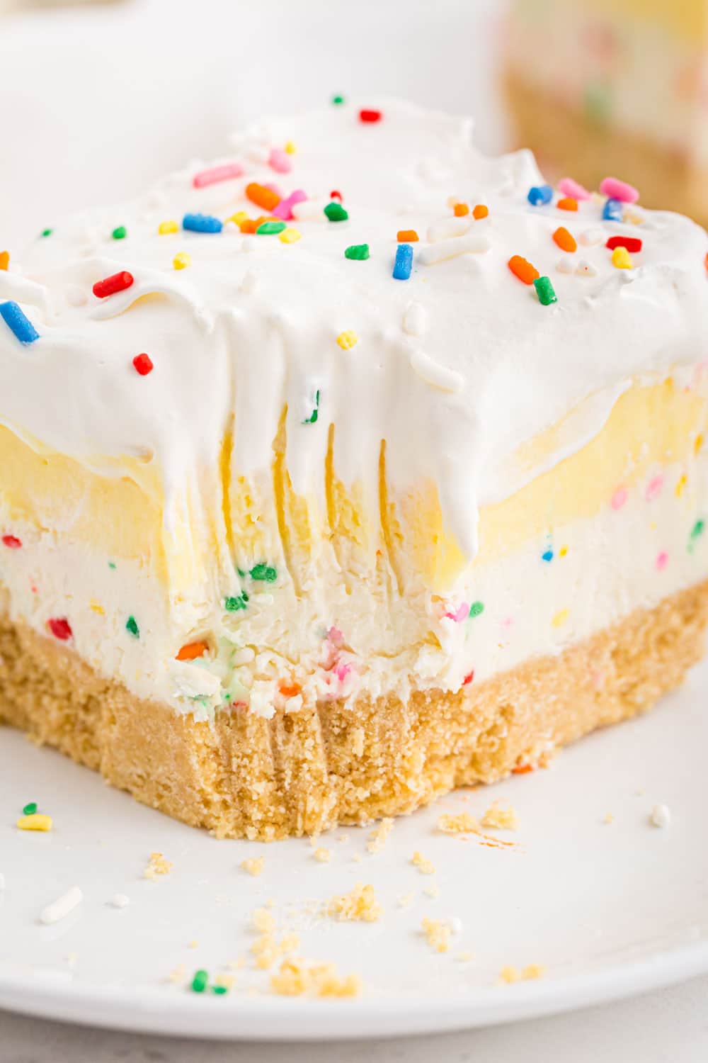 Slice of birthday cake cheesecake bars on a white serving place with a forkful of dessert removed from the dessert bar, placed on a white marble surface. 