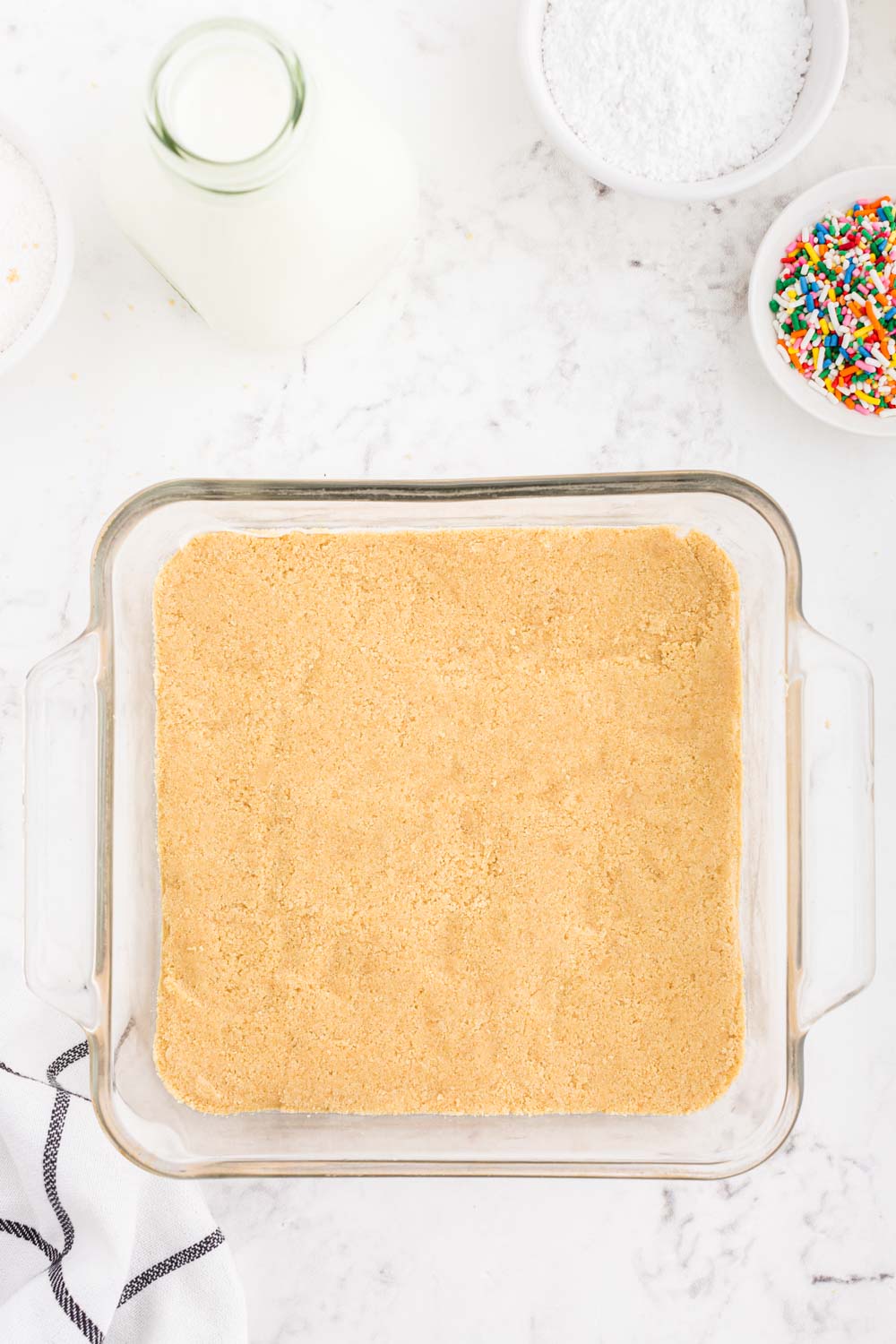 Vanilla cookie base pressed into a 8x8-inch glass dish, small bowl of rainbowl sprinkles, glass jar of whole milk, bowl of powdered sugar, striped linen, on a white marble surface.