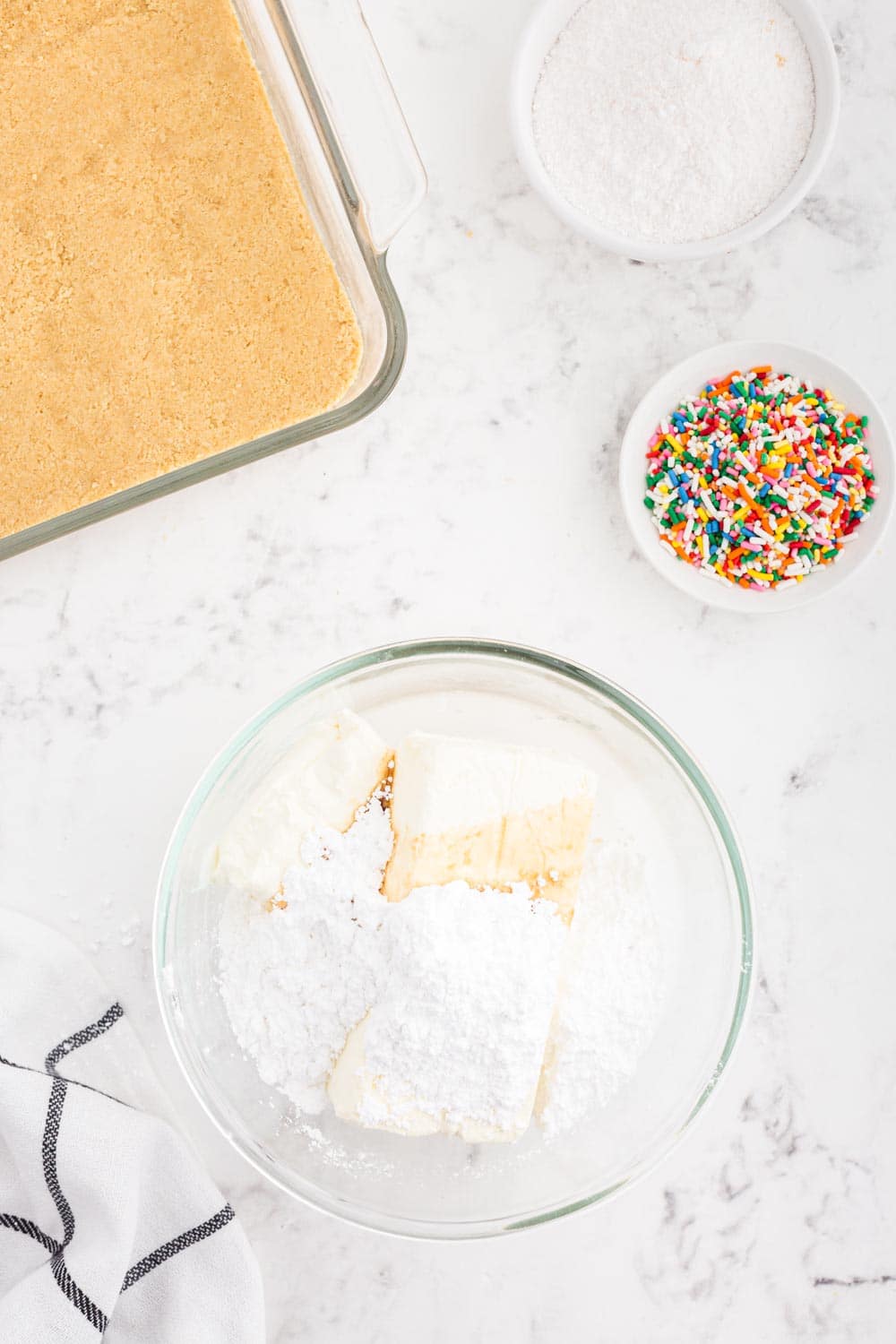 Cream cheese, powdered sugar, vanilla extract, and whipped topping added to a standing mixer bowl, small bowl of rainbow sprinkles, bowl of powdered sugar, glass dish with cookie crust, striped linen, on a white marble surface.