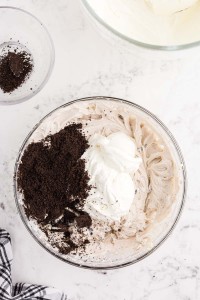 Crushed Oreo cookies and whipped cream added to the cream cheese mixture, checked linen, bowl of remaining crushed Oreos, bowl of whipped cream, on a white marble countertop