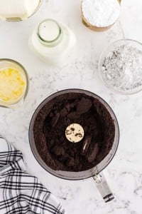 Crushed Oreo cookies in a food blender, bowl of melted butter, jar of heavy whipping cream, checked linen, bowl of powdered sugar, bowl of pudding mix, on a marble countertop