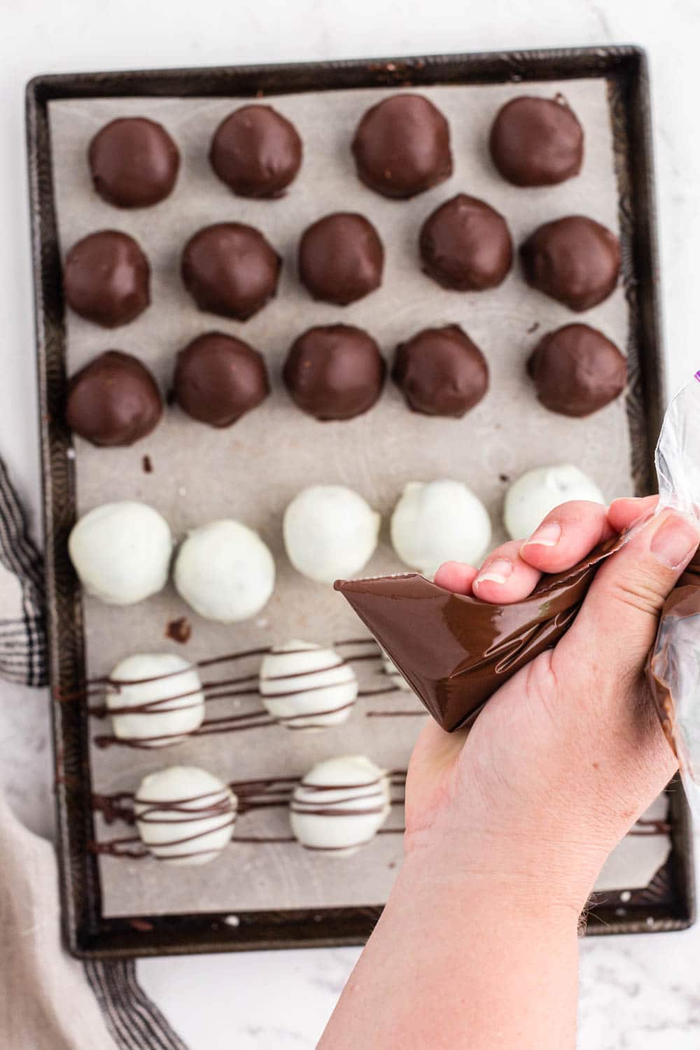 Chocolate coated Oreo balls, melted dark chocolate in piping bag drizzled over white chocolate coated Oreo balls on parchment paper-lined tray, beige linen, on a white marble countertop