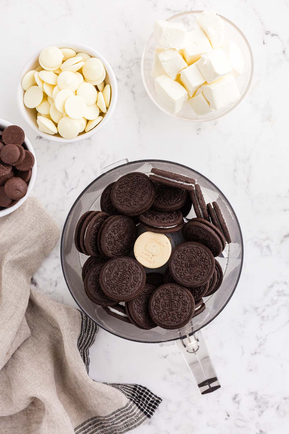 Whole Oreo cookies in a food processor, bowl of cream cheese, bowl of dark chocolate melting wafers, bowl of white chocolate melting wafers, beige linen, on a white marble countertop