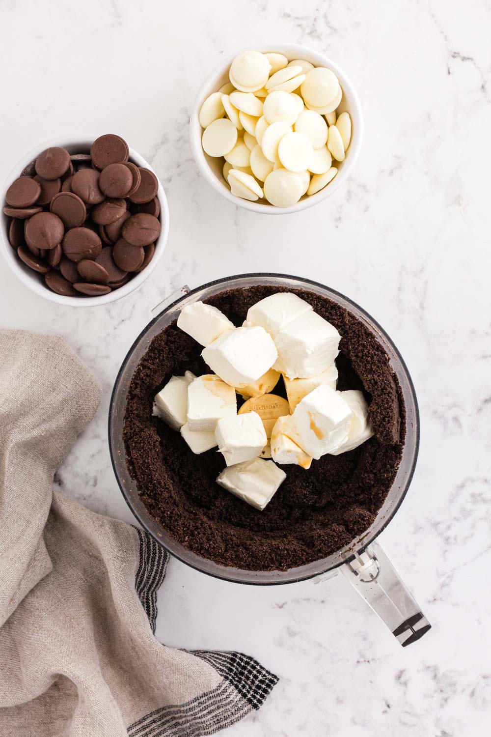 Cream cheese and vanilla extract added to crushed Oreo cookies in food processor, bowl of dark chocolate melting wafers, bowl of white chocolate melting wafers, beige linen, on a white marble surface