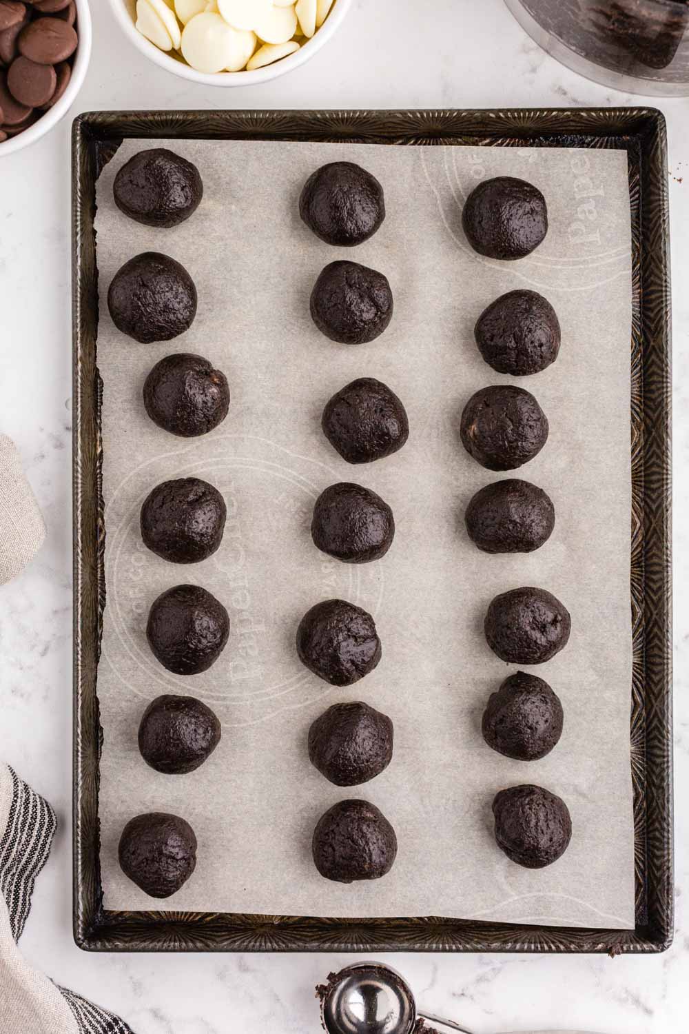 Parchment paper-lined baking sheet with frozen Oreo balls, metal cookie scoop, bowl of dark chocolate melting wafers, bowl of white chocolate melting wafers, beige linen, on a white marble countertop