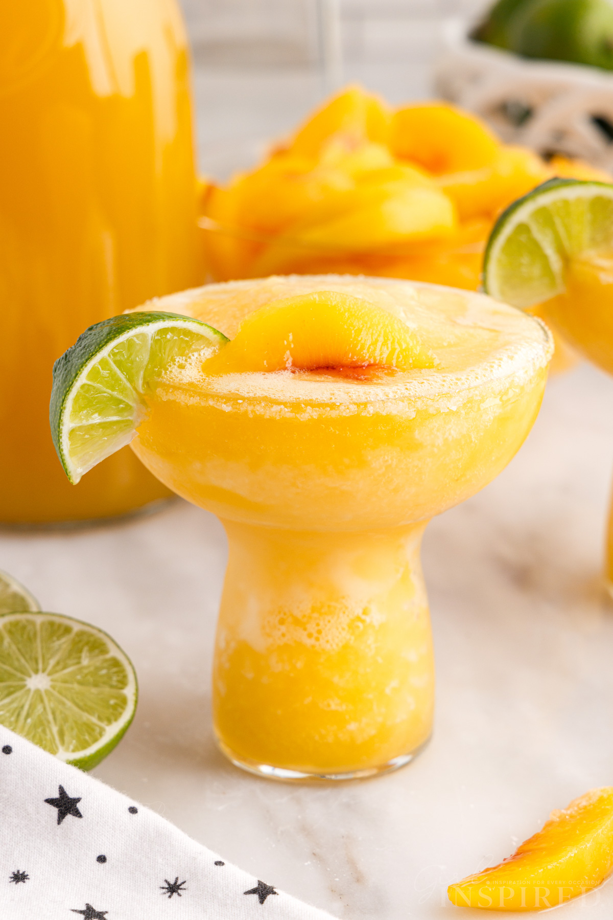 frozen peach margarita garnished with a lime wedge, bowl of peaches, and jar of peach juice.