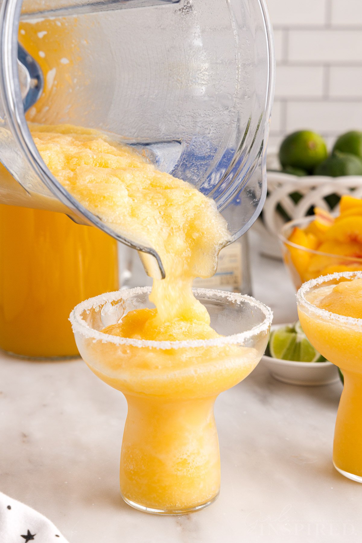 Peach margarita pouring from blender into prepared glass, bowl of limes and bowl of peach slices.