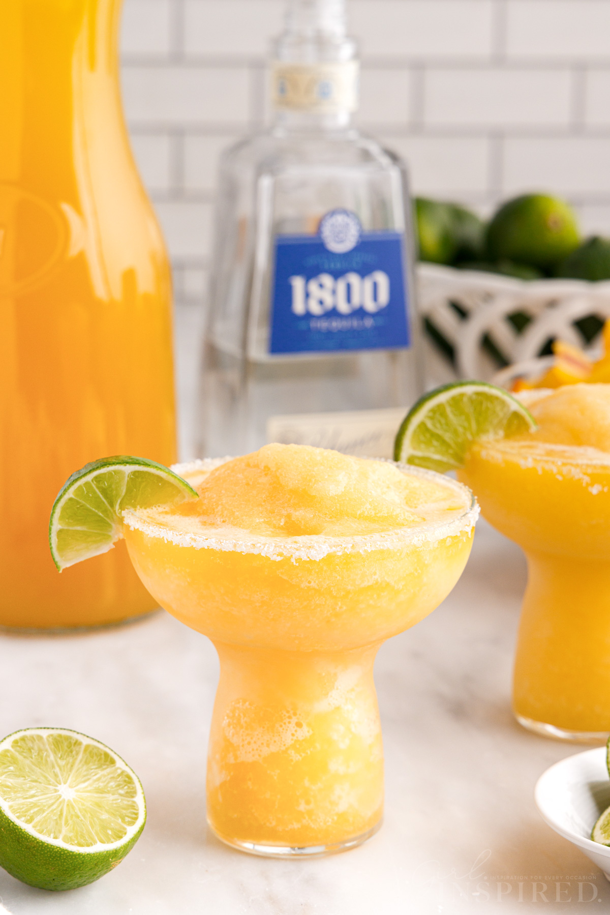 Frozen peach margaritas mounded in margarita glasses with lime wedges, bottle of 1800 tequila, jar of peach juice, bowl of limes.