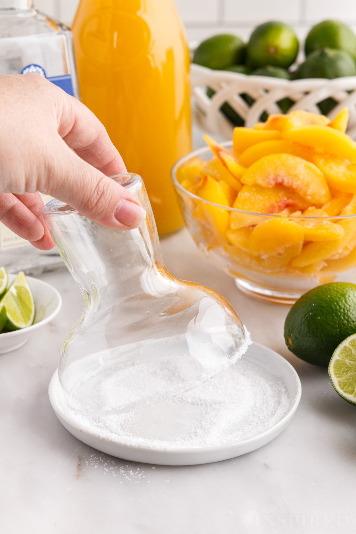 Tipping margarita glass into dish of salt to salt the rim, bowl of peaches, lime wedges.