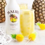 Pineapple Spears in a glass of Malibu Rum next to the bottle and paper pineapples on toothpicks