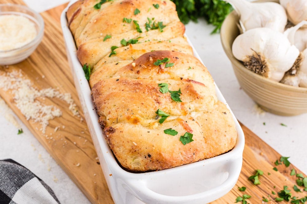 Pull apart garlic bread in a white bread loaf baking dish on top of a wooden kitchen board, bowl of whole garlic cloves, fresh parsley, bowl of grated parmesan cheese, black and white checkered linen, on a white marble surface.