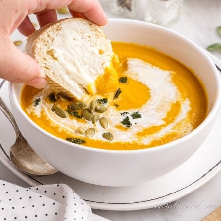 Close up of dipping a piece of bread into pumpkin soup topped with pepitas