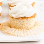 Close up of single white cupcake with vanilla frosting, cupcake liner removed from the cupcake, additional cupcakes on a white platter, on top of a white marble countertop