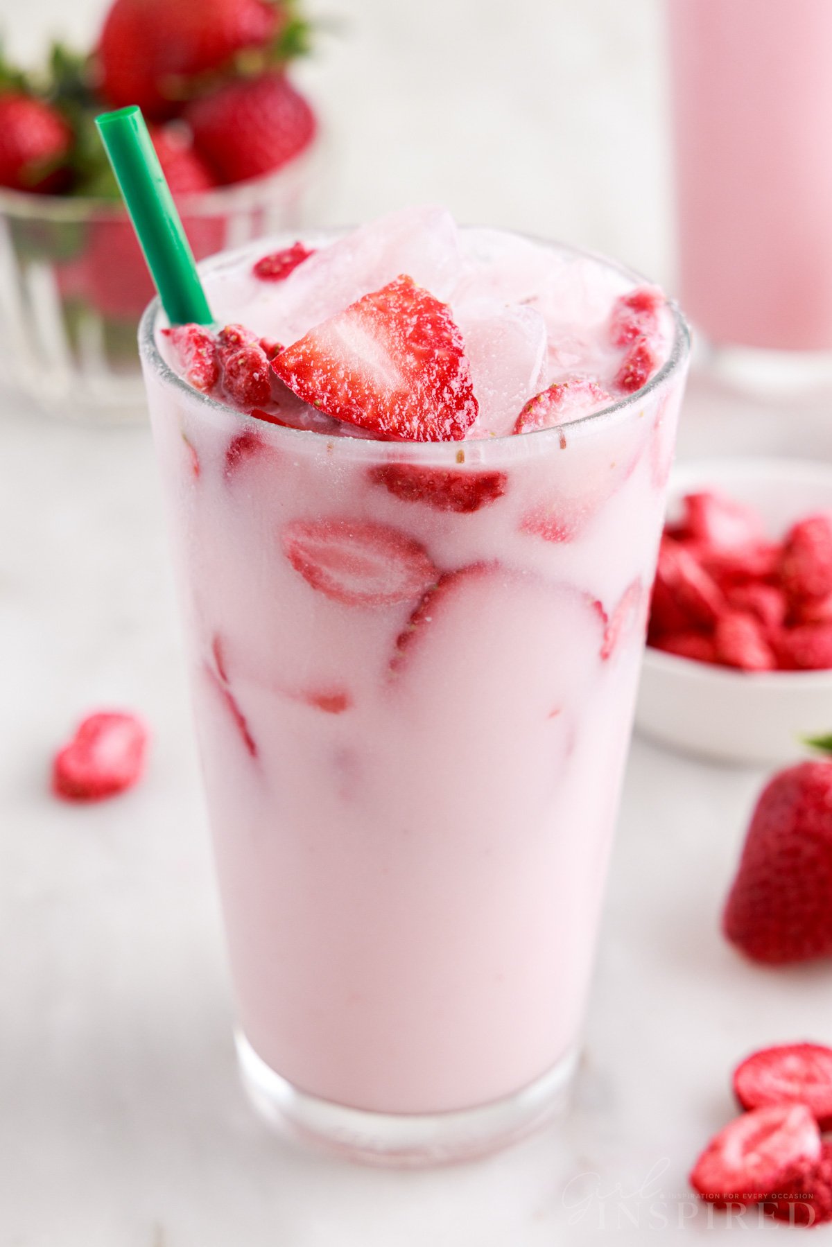 Pink drink in glass with straw and fresh strawberry slices on top, bowl of strawberries.