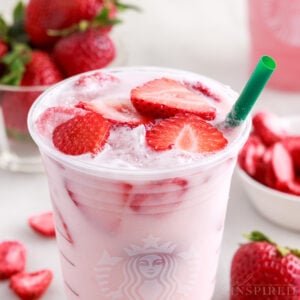 Pink drink recipe filling a plastic cup with fresh strawberries floating on top, bowl of strawberries, green straw.