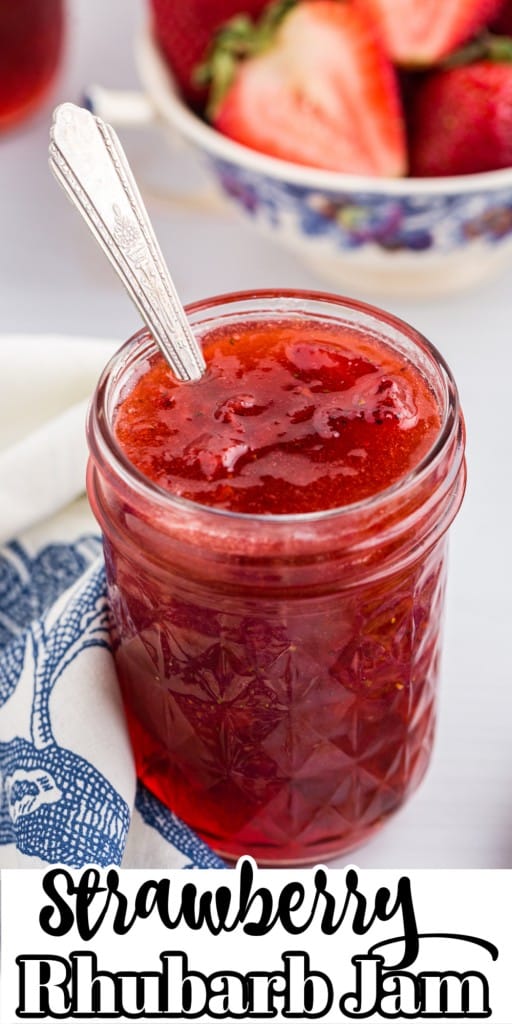 Close up of a glass jar of strawberry rhubarb jam with a bowl of cut strawberries in the background