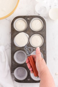Cupcake mixture spooned into cupcake liners in a muffin tray, bowl of cupcake batter, white linen, on a white marble countertop