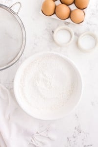 Bowl of sifted flour, bowl of baking powder, bowl of salt, sieve, tray of eggs, on white marble countertop