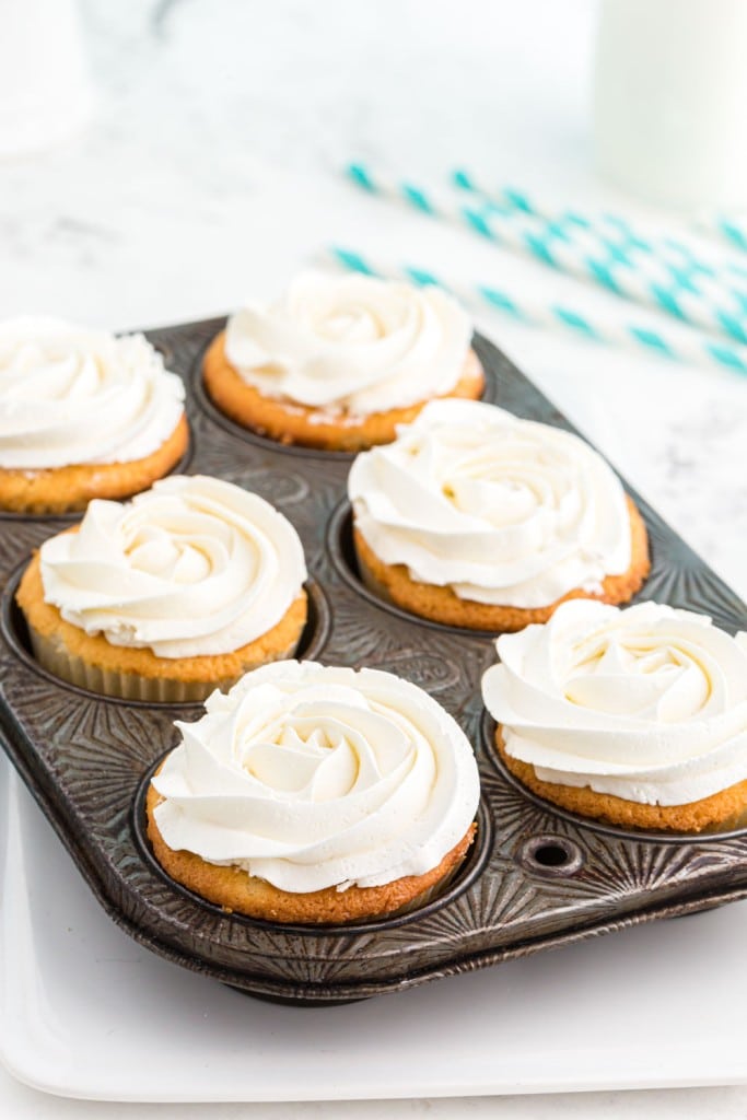 Muffin tray filled with six baked vanilla cupcakes with frosting on a white plate, blue and white striped paper straws, on white marble countertop